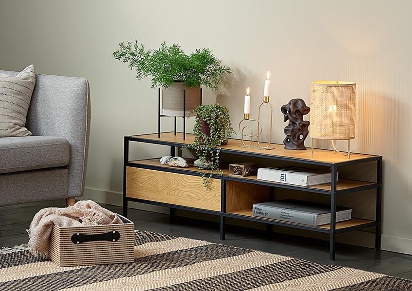 TRAPPEDAL TV stand, ALBERT table lamp, STEN ornament, ERVIN candlestick, FREJ basket A corner of a living room with a sofa, a basket, a small table lamp and a modern TV stand